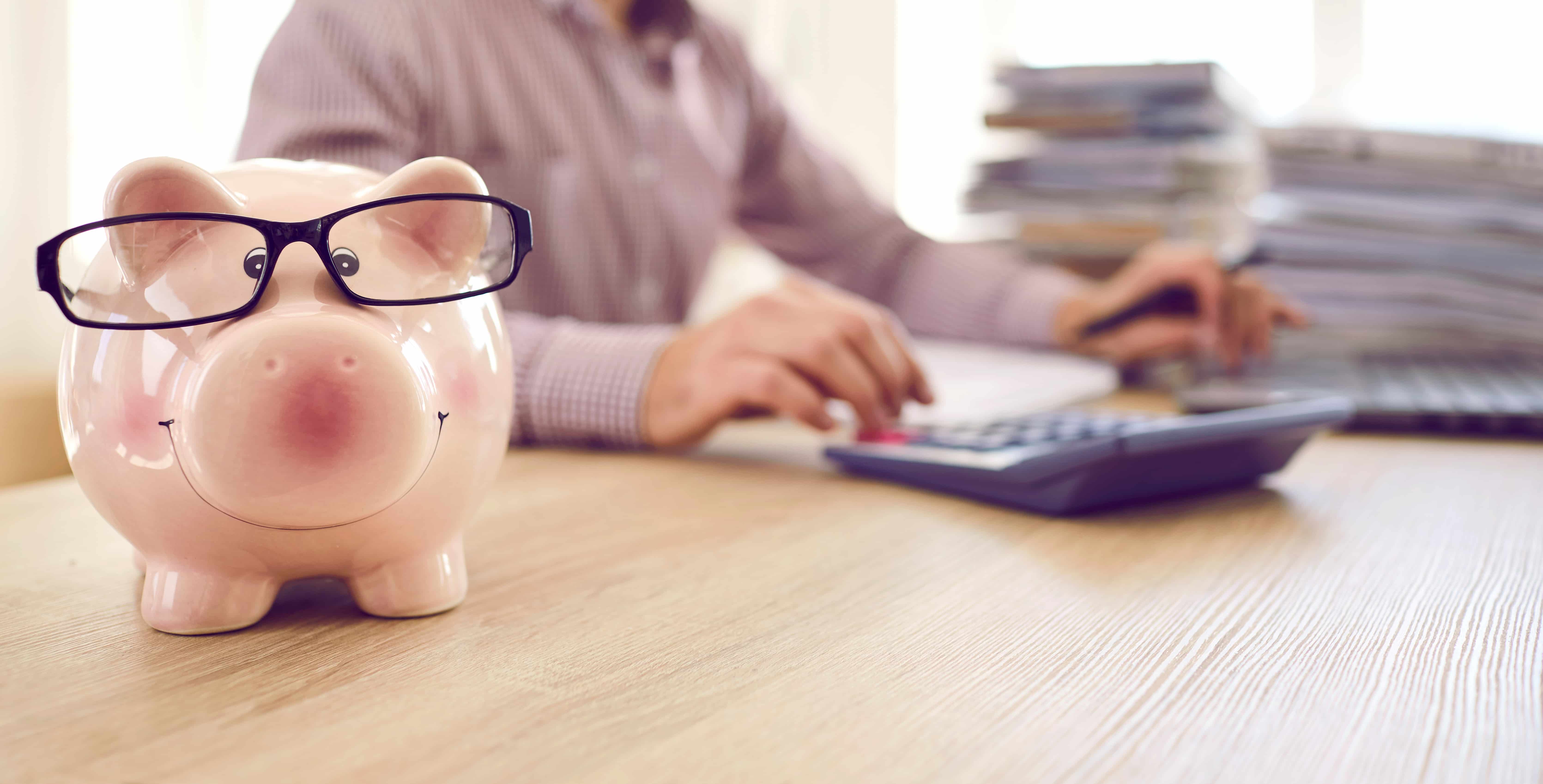 Banner with close up of cute piggy bank wearing eyeglasses placed on table of accountant who's working with tax documentation, analyzing budget and using calculator and laptop computer in background